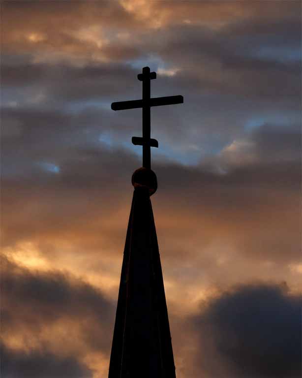Kirchturm mit Kreuz vor dramatischem Abendhimmel.
