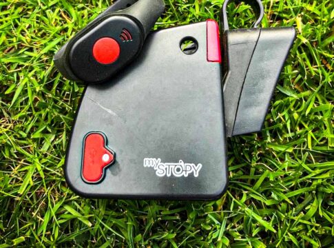 Zu sehen ist das mySTOPY Bremsmodul mit schwarzer Armband-Fernbedienung und rotem Knopf, liegend auf grünem Rasen. Es handelt sich um einen Stopper fürs Kinderlaufrad zur Fernsteuerung.