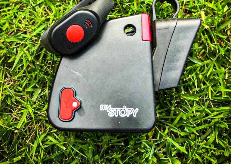 Zu sehen ist das mySTOPY Bremsmodul mit schwarzer Armband-Fernbedienung und rotem Knopf, liegend auf grünem Rasen. Es handelt sich um einen Stopper fürs Kinderlaufrad zur Fernsteuerung.