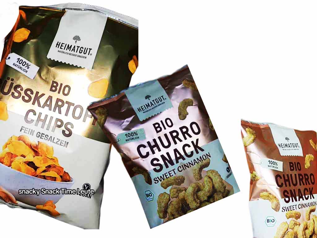 Zu sehen sind drei verschiedene Snackverpackungen der Marke Heimatgut nebeneinander. Links: Bio-Süßkartoffelchips, fein gesalzen. In der Mitte und rechts daneben: Bio-Churro-Snack in der Sorte „Sweet Cinnamon”. Auf allen Tüten steht „100 % natürlich“. Die farbenfrohen Produkte richten sich an bewusste Snack-Fans.