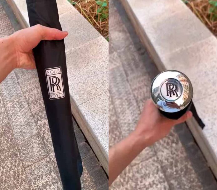 Rolls-Royce-Regenschirm mit silbernem RR-Logo am Griff und in einer Hülle, die von einer Hand auf einem Steinboden gehalten wird.